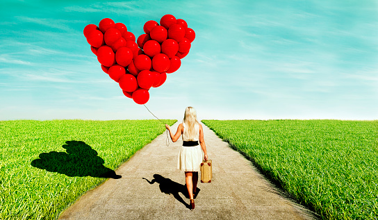 Female wearing a dress holds on to a suitcase and a string with red balloons attached. The balloons forms a heart. The woman walks on a path towards the horizon. Concept of looking for love.