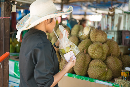 In Phuket, Thailand a woman holds a fresh cut coconut in a plastic bag as she drinks its water through a straw and holds it with baht in her hand. A stack of durian is visible in the background as she walks through the street market.