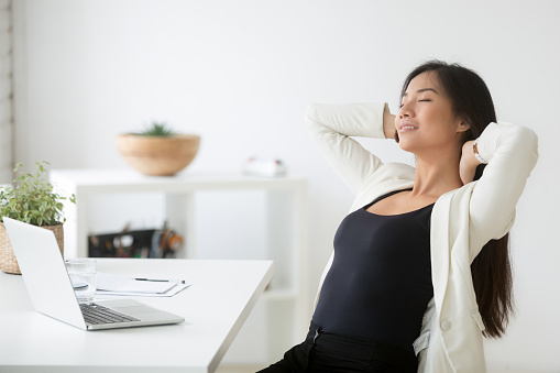 Relaxed happy asian woman enjoying break at workplace, calm smiling korean employee relaxing from computer holding hands behind head breathing air, positive thinking, feeling no stress free at work