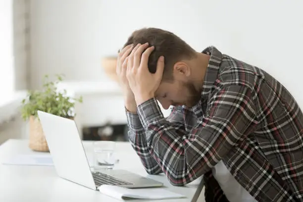 Frustrated depressed man holding head in hands shocked by bankruptcy stock downfall sitting at work desk with laptop, stressed tired businessman feels despair lost money online or got problem debt