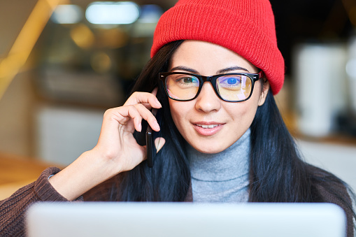Portrait of beautiful trendy woman wearing glasses speaking by phone and  using laptop while working in modern co-working space or cafe