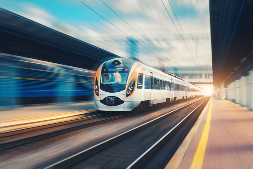 Speed passenger train in motion on the railway station at sunset in Europe. Modern intercity train on railway platform with motion blur effect. Urban scene with railroad. Railway transportation