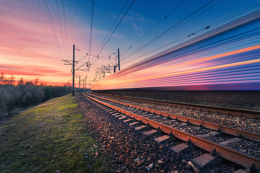High speed passenger train in motion on railroad at sunset. Blurred modern commuter train. Railway station and colorful sky. Railroad travel, railway tourism. Industrial landscape. Transportation