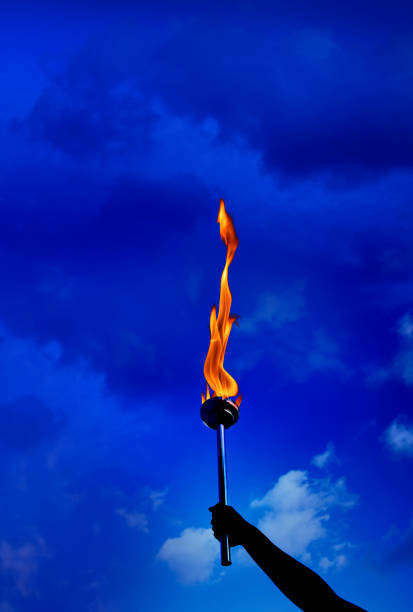 istock Close up hand holding burning flaming torch with flames over cloudy blue sky 953256504