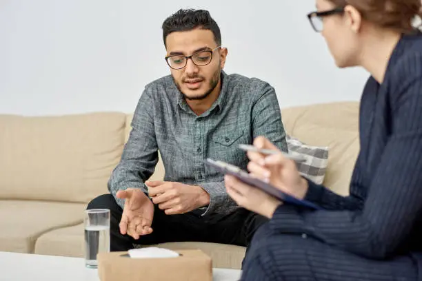 Young mixed-race man wearing eyeglasses sitting on cozy sofa and discussing faced problem with highly professional psychologist during therapy session at cozy office