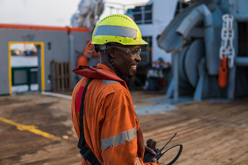 Seaman AB or Bosun on deck of offshore vessel or ship , wearing PPE personal protective equipment - helmet, coverall, lifejacket, goggles. He holds VHF walkie-talkie radio in hands.