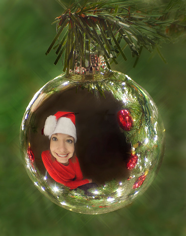 A picture of a girl’s reflection in a silver Christmas ornament hanging on a Christmas tree.