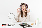 Fun crazy dizzy loony wild mad woman with curlers on hair sitting at table applying makeup with set facial decorative cosmetics isolated on white background. Beauty female fashion lifestyle concept.