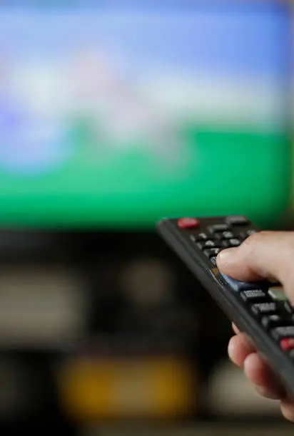 A hand holding a remote control with a TV in the background
