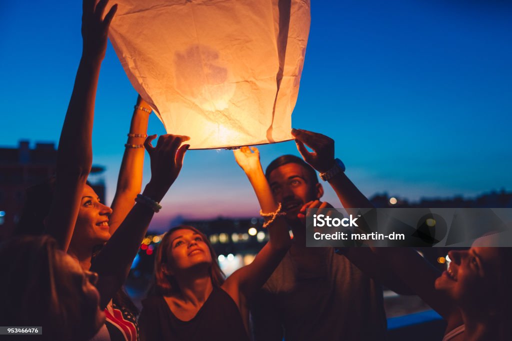 Friends on rooftop party releasing paper lantern Happy friends holding a burning lantern ready to fly in the sky Hot Air Balloon Stock Photo