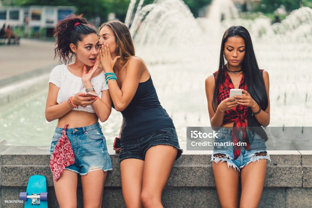 Young woman telling a secret to a friend Girls hanging outside in the city park and gossiping Gossip Stock Photo