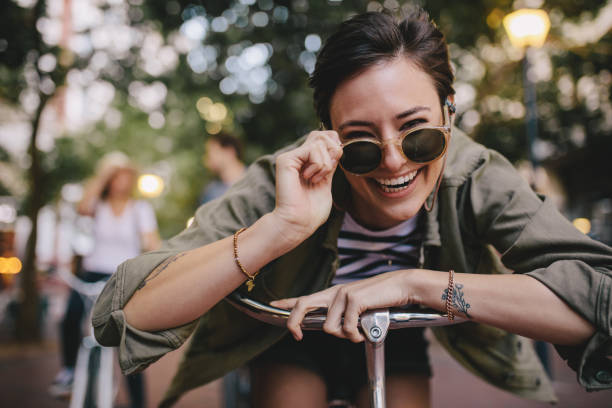 Cheerful woman in sunglasses with bike Cheerful woman wearing sunglasses leaning on her bike outdoors. Woman enjoying outdoors with friends at the back. hipster bike stock pictures, royalty-free photos & images