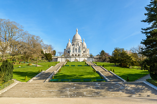 Front view of the bright white basilica of the Sacred Heart of Paris, situated at the top of the Montmartre hill, from the bottom of the stairways in the Louise Michel park by a sunny spring morning.