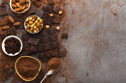 Dark broken chocolate pieces on dark background, cocoa powder and various nuts in bowls closeup. Sweet wallpaper, confectionery shop advertising and cooking ingredients concept, topv iew, copy space