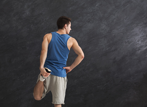 Young man warming up before training at gym. Guy stretching his leg at gray background indoors, copy space, back view