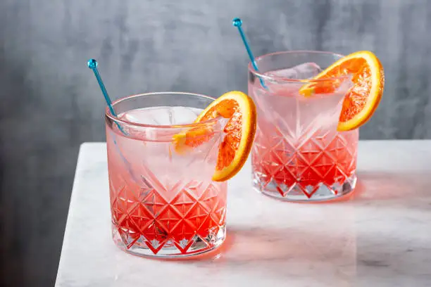Two cold gin and tonics made with blood orange syrup and garnished with blood orange slices. The cocktails are served on ice with blue stirrers. On a minimalist marble bar counter with a gray background with copy space.
