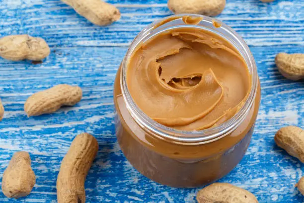 peanut butter in an open jar on a blue wooden background, next to peanuts scattered