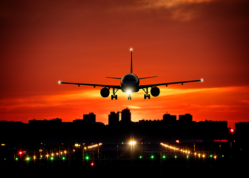 Passengers commercial airplane landing on runway with dramatic sunset sky. Modern cityscape with silhouettes of skyscrapers on background. Concept of fast travel