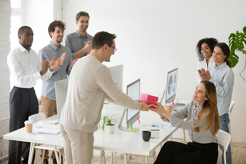 Friendly diverse corporate team congratulating happy female colleague with birthday and applauding, male employee presenting gift box greeting woman making pleasant surprise to coworker in office