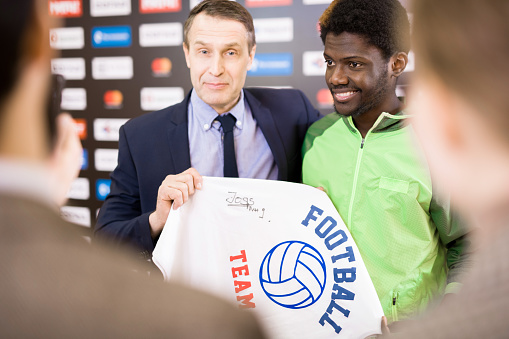 Portrait of smiling African-American sportsman taking selfie with fan posing for photo with his coach and holding signed t-shirt