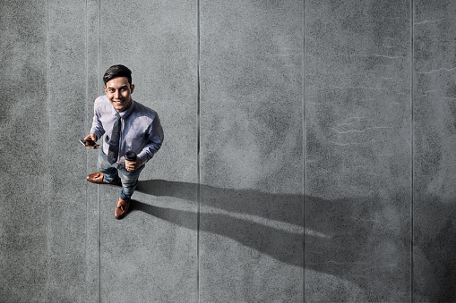 Happy Young Man Standing on Concrete Floor, Smiling and Looking at Camera. Hand holding Mobile Phone and Coffee. Top View