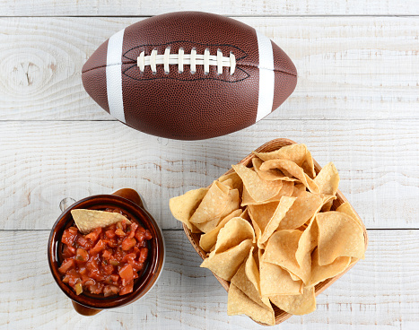 High angle shot of a bowl of corn chips a crock full of fresh salsa and an American football on a whitewashed rustic wood table. Horizontal format.