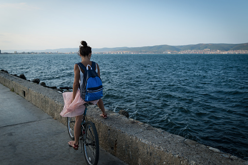 Young stylish woman in dress and with backpack is riding bicycle on promenade on background of sea, city and hills afar.
