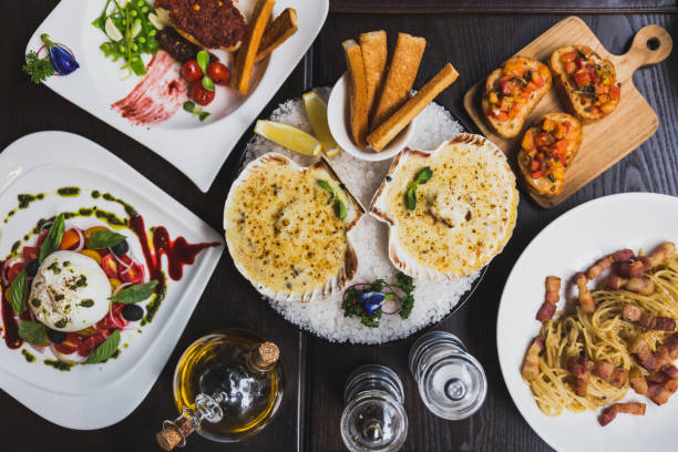 istock Italian dishes on the table including Burrata Salad, Baked Cod with sauce,  Spaghetti Carbonara, Baked scallops with cheese and Canapes with olive oil, pepper and salt on the table. 953704836