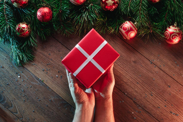 istock A frame of pine branches and Christmas decorations and hands giving a gift on the background of a wooden table. Merry Christmas and happy holidays. View from above. 953724652