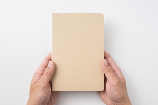 Design concept - Top view of man's hand holding hardcover kraft notebook isolated on white background for mockup