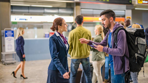 Backpacker at the airport Young male backpacker asking young smiling female airport attendant about ticket for his flight, incidental people in background. check in person stock pictures, royalty-free photos & images