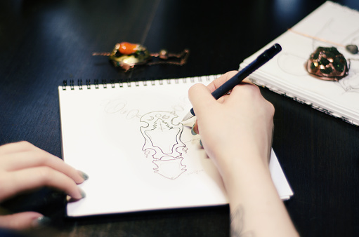 Artist designer girl draws a sketch of unusual jewelry ornaments with a pen on white paper. Home workshop