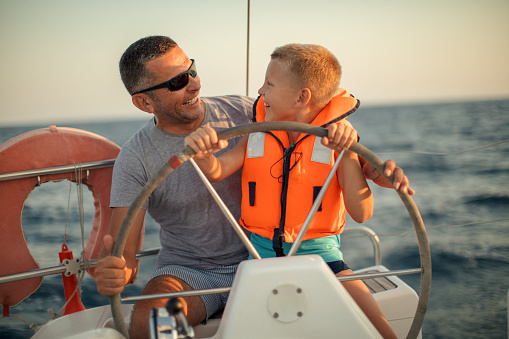 One little boy on sailboat sitting with his father, enjoying at summer and having a fun while steering with yacht at sunset.