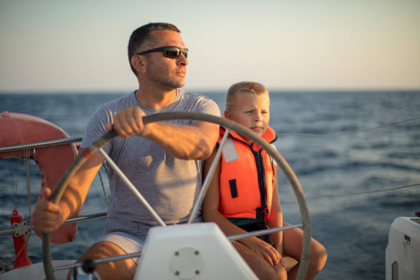 istock Little boy sitting with father who steering with sailboat 953779400