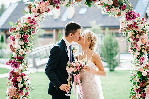 The bride and groom kissing. Newlyweds with a wedding bouquet, holding glasses of champagne standing on wedding ceremony under the arch decorated with flowers and greenery of the outdoor.