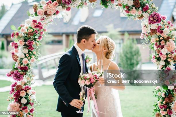 The Bride And Groom Kissing Newlyweds With A Wedding Bouquet Holding Glasses Of Champagne Standing On Wedding Ceremony Under The Arch Decorated With Flowers And Greenery Of The Outdoor Stock Photo - Download Image Now