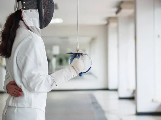 Close-up of a fencer in white fencing suit and holding a sword on gym background.sport concept.