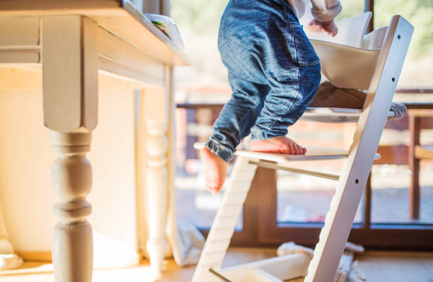 Toddler boy in a dangerous at home, climbing into highchair. Little toddler boy climbing on wooden highchair at home. Domestic accident. Dangerous situation at home. high chair stock pictures, royalty-free photos & images