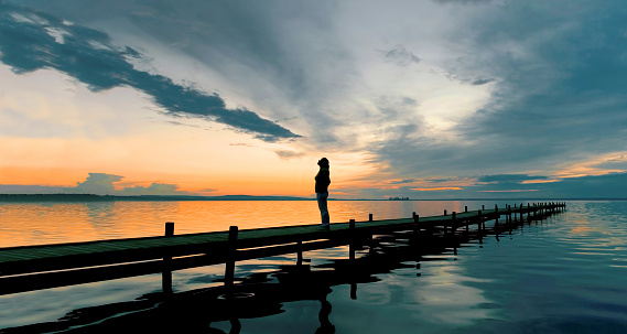 Silhouette on woman standing on jetty watching dramatic orange glow of sky after sunset