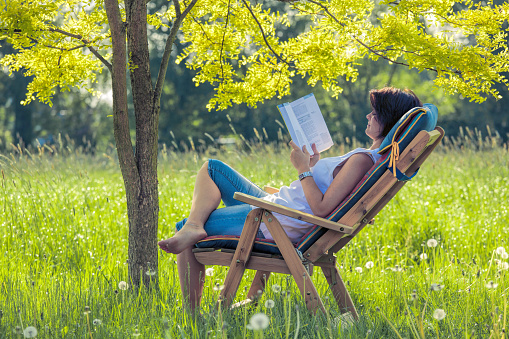 Young woman lying on deck chair in shadow under tree reading book