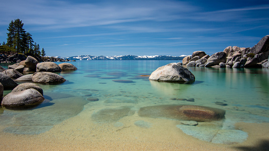 Long exposure of Sand Harbor at Lake Tahoe North, Nevada county, California, USA, featuring blue transparent water, rocky shore and snow on tops of background mountains on a blue sky day with few clouds- travel California