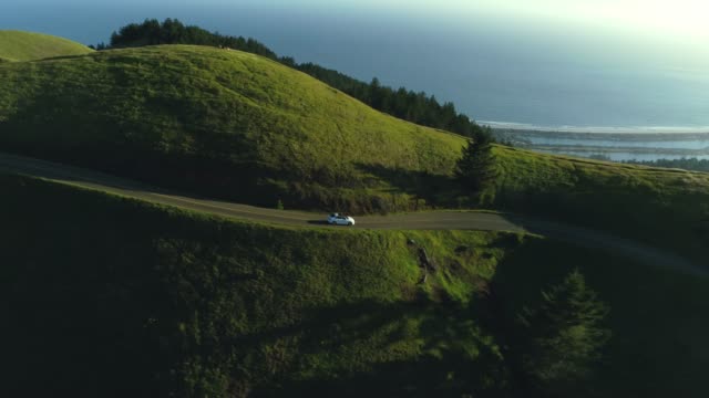 Aerial view of car driving down country road through rural rolling hills with ocean in background at sunset
