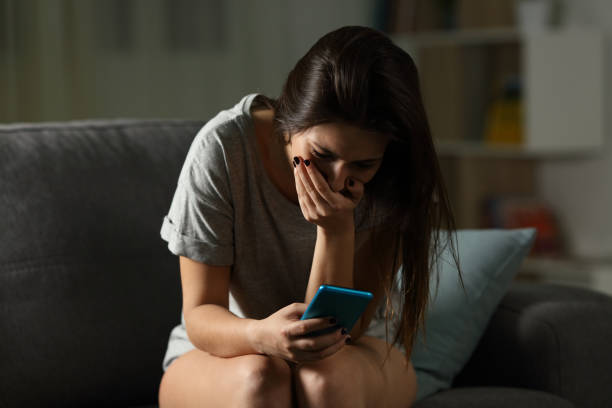 Sad teen receiving bad news online at home Sad teen receiving bad news online in a smart phone sitting on a couch in the living room at home harassment stock pictures, royalty-free photos & images