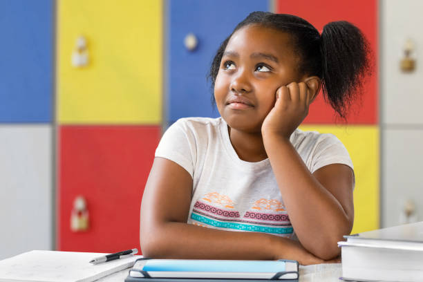 istock Little african student sitting at desk in classroom wondering. 953900740