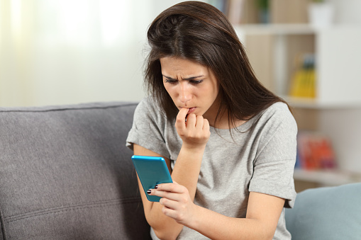 Nervous girl biting nails reading phone content sitting on a couch in the living room at home