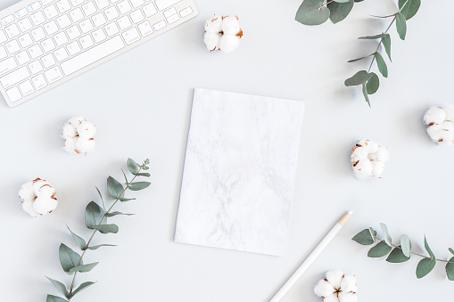 Workspace with computer, marble paper blank, cotton flowers and eucalyptus branches on pastel blue background. Flat lay, top view, copy space