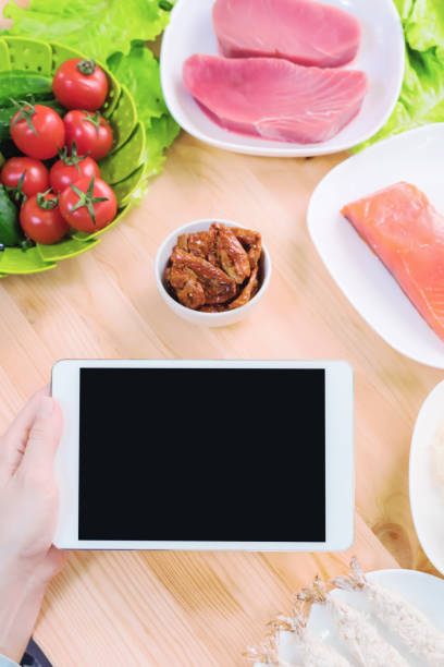 istock Close-up top view of female hands holding a tablet computer with a blank display surrounded by healthy food on a home kitchen table. Workpiece to insert the design of the mobile application into the screen. Products of Greek cuisine raw seafood and fresh 953945870