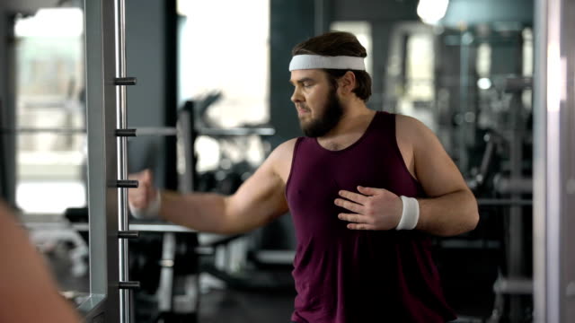 Funny fat man looking at mirror reflection gym and posing, pretending muscular