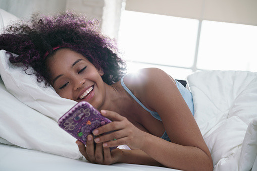 Young African American woman in bed at home. Happy black girl smiling while texting message on mobile telephone. Latina teenager lying in bed and using cell phone for social media and internet. Slow motion