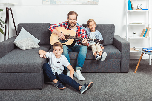father with guitar and cute little sons smiling at camera at home